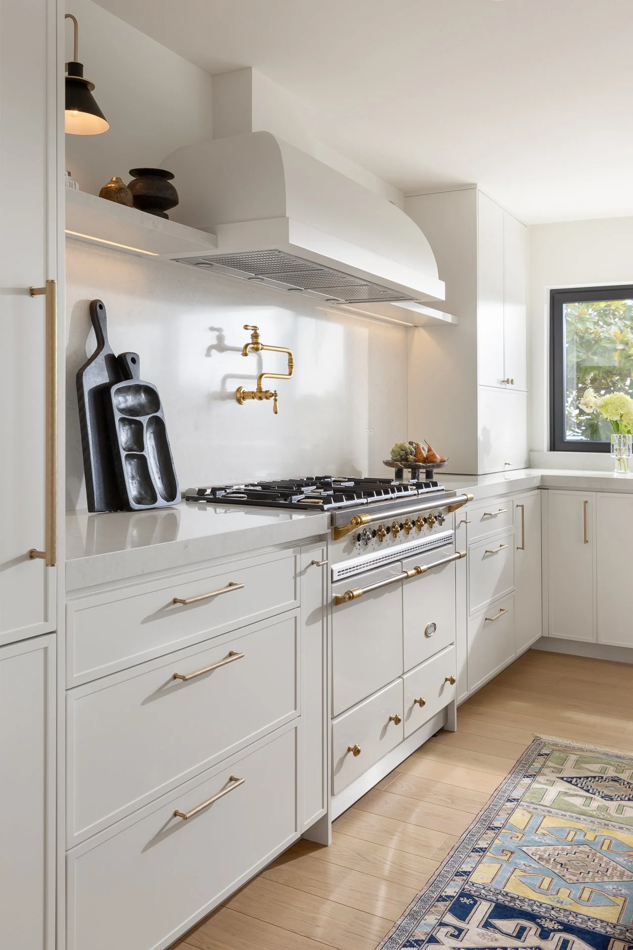 White kitchen with gold cabinet handles, a gas stove with gold handles, black cutting boards, a brass faucet, a window with a vase of white flowers, and a patterned rug on wooden flooring.