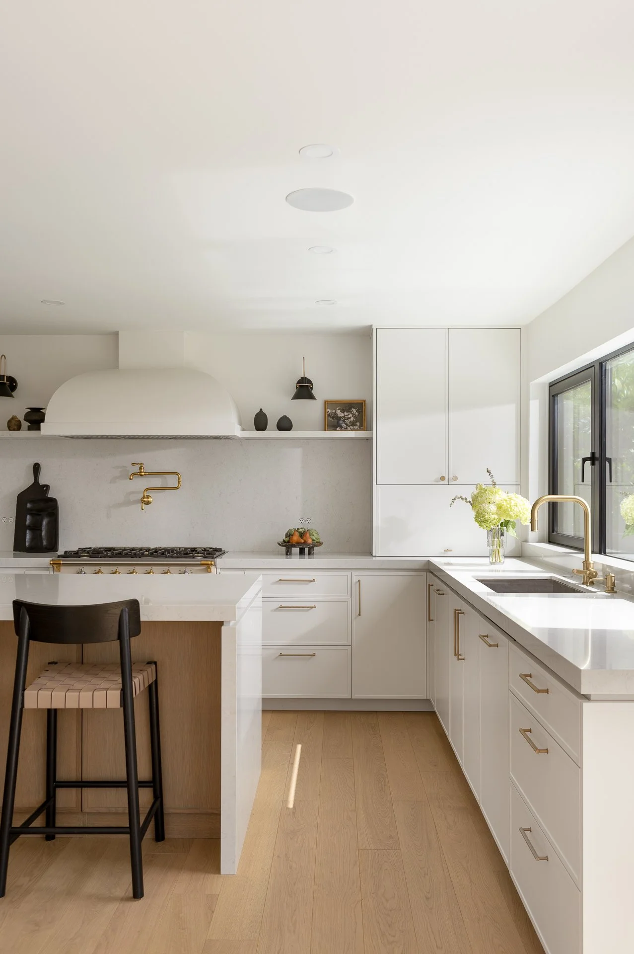 Modern kitchen with white cabinets, gold hardware, a gold faucet, and a large window with a vase of flowers on the counter.