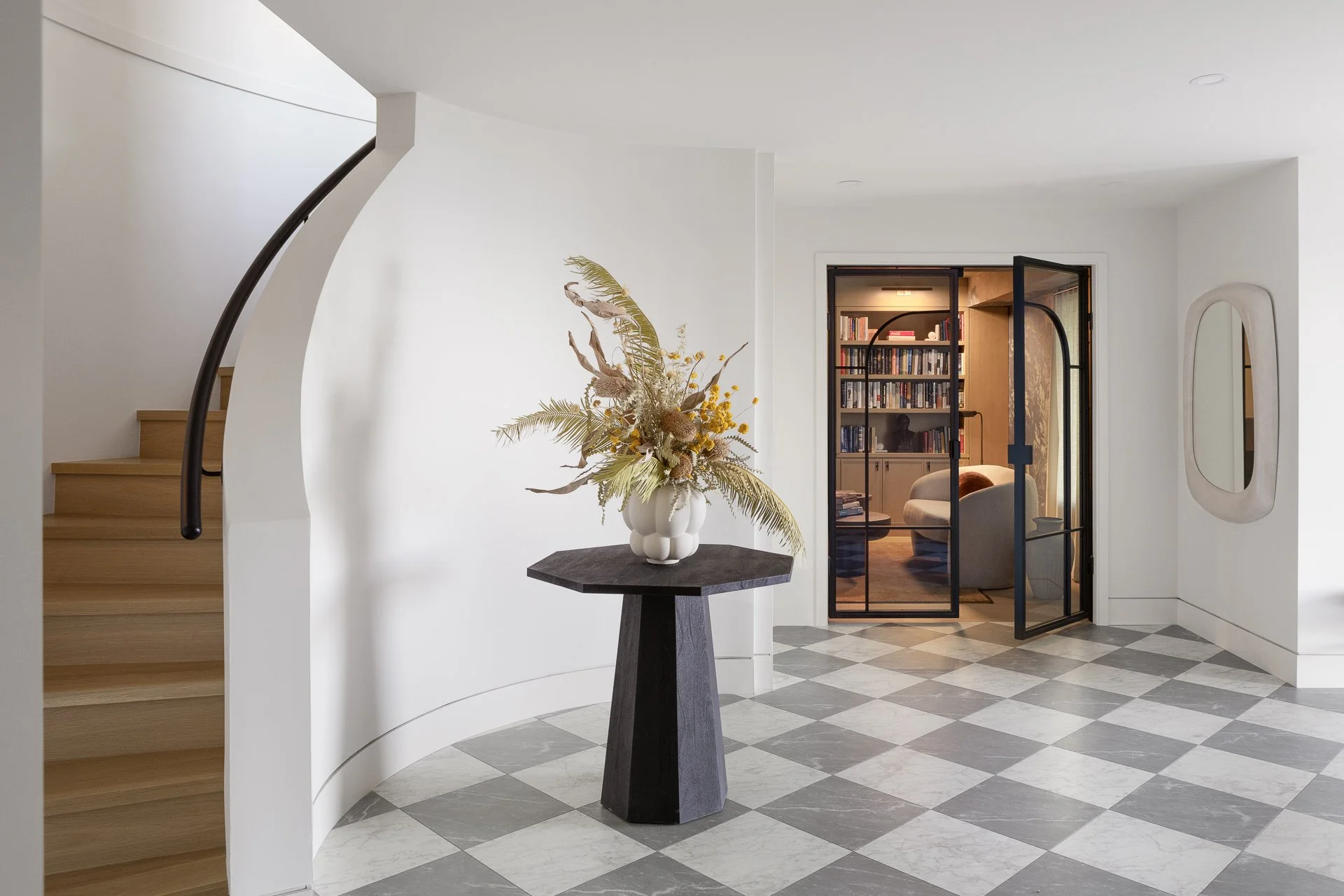 Interior of a modern home with a black table holding a white vase with dried flowers, a staircase on the left, and a room with bookshelves and seating visible through a glass door on the right.