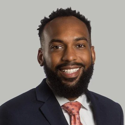 Headshot of a man with dark skin, curly hair, and a beard, wearing a navy suit, white shirt, and pink tie, smiling against a gray background.