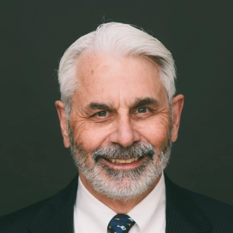 Close-up portrait of a senior man with gray hair and beard, smiling, wearing a dark suit, white shirt, and dark tie with small light-colored patterns, against a dark background.