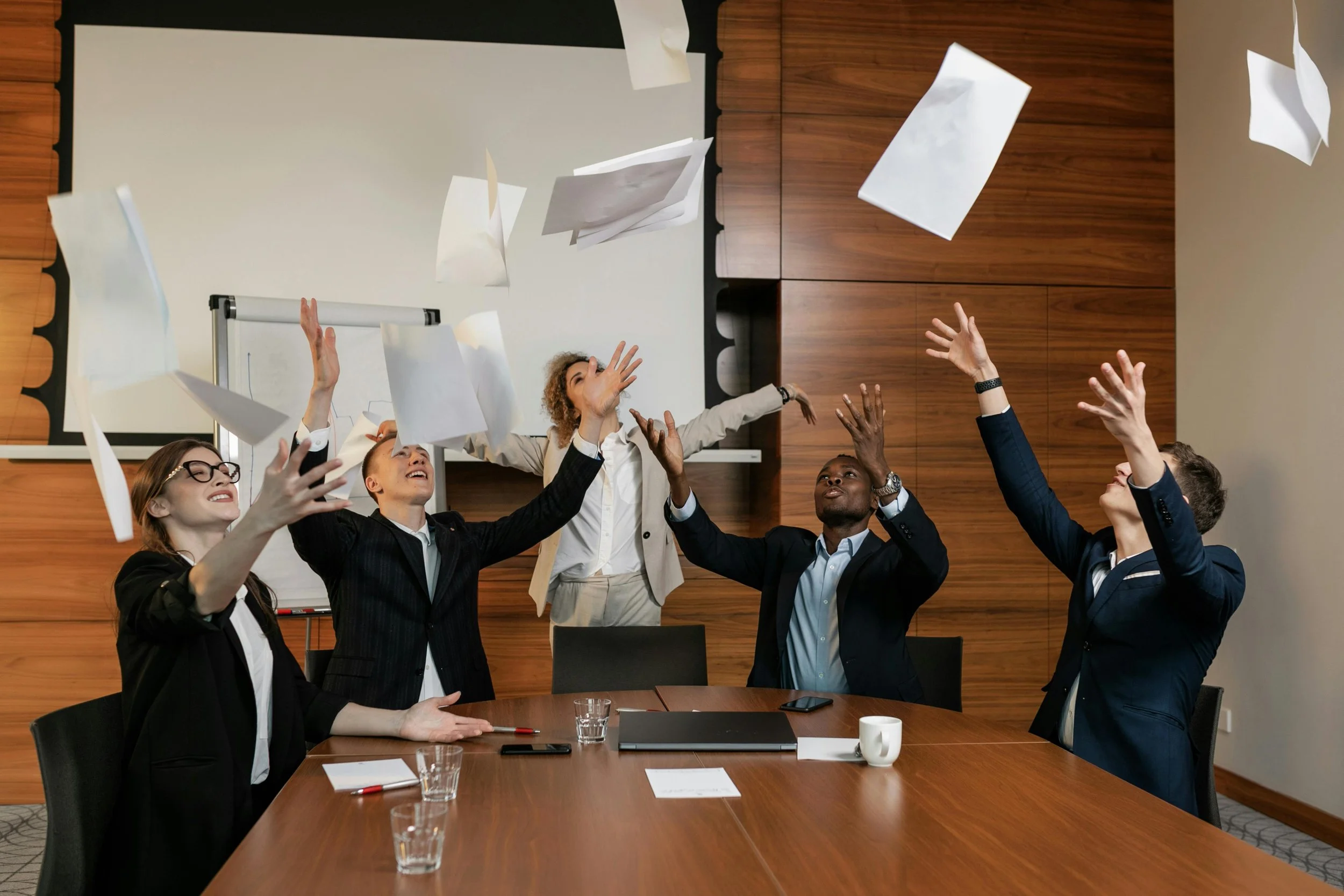 A group of business people are throwing up papers happily in a boardroom.