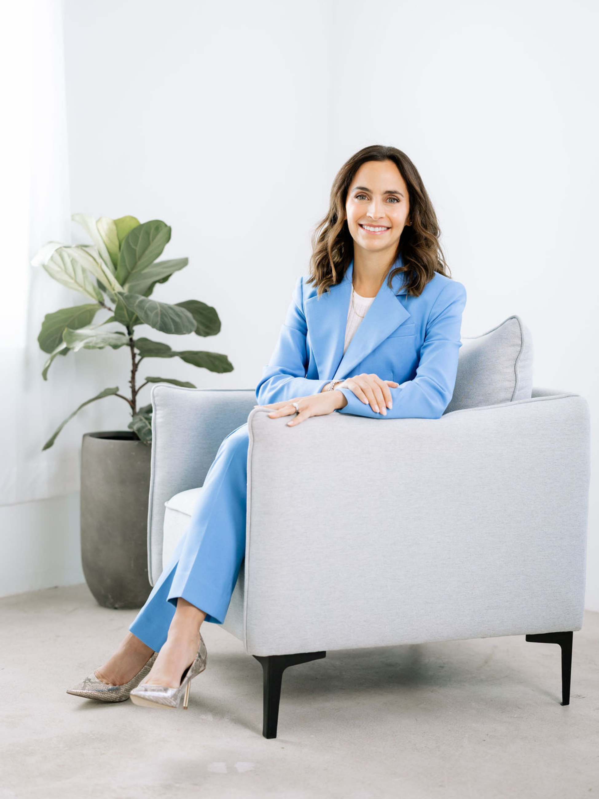 A brunette woman wearing a blue suit is sitting cross legged and smiling in a chair.