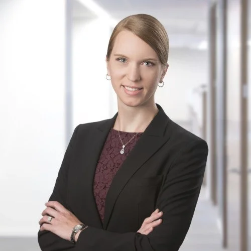 A professional woman with short brown hair, wearing a black blazer and a maroon lace top, standing in a modern office hallway with a smiling expression.