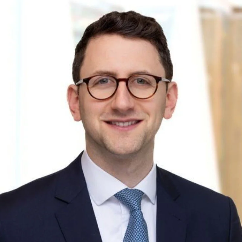 A young man wearing glasses, a dark suit, and a blue tie, smiling in a professional setting.