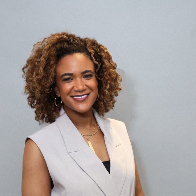A smiling woman with curly hair wearing a sleeveless light-colored blazer and gold jewelry standing against a light gray background.