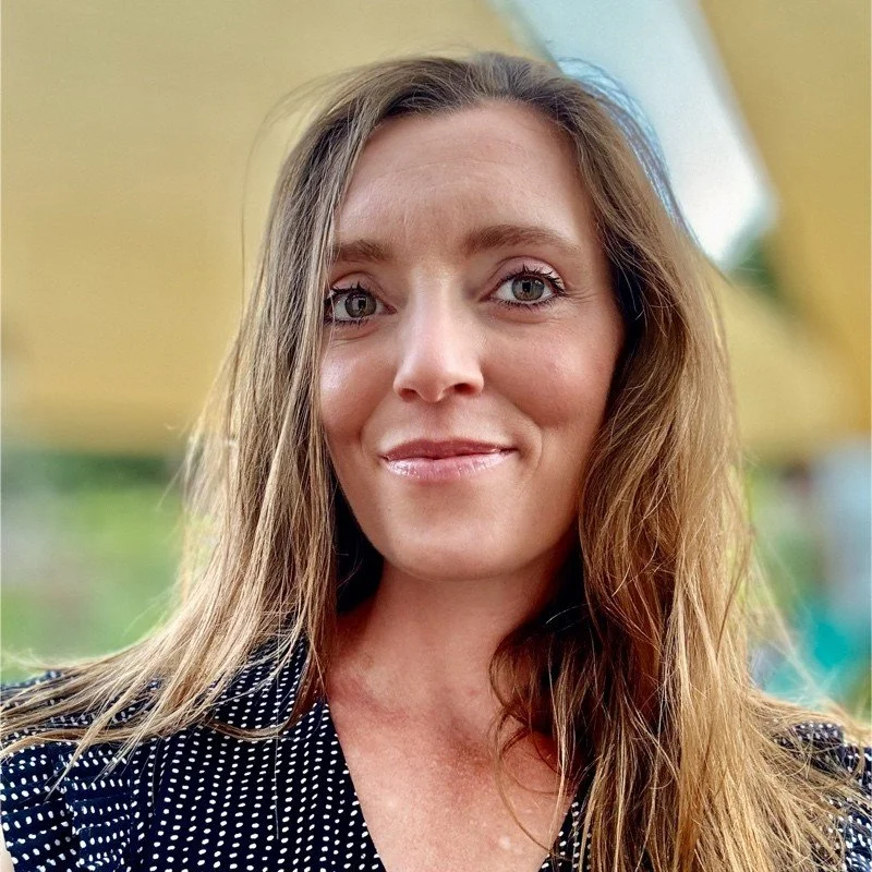 Close-up of a woman with long, wavy brown hair wearing a black and white polka-dot top, smiling softly outdoors.