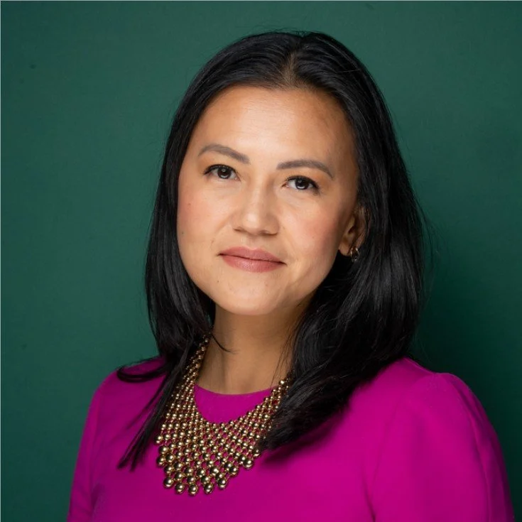 A woman with long black hair wearing a pink top and a layered gold necklace against a green background.