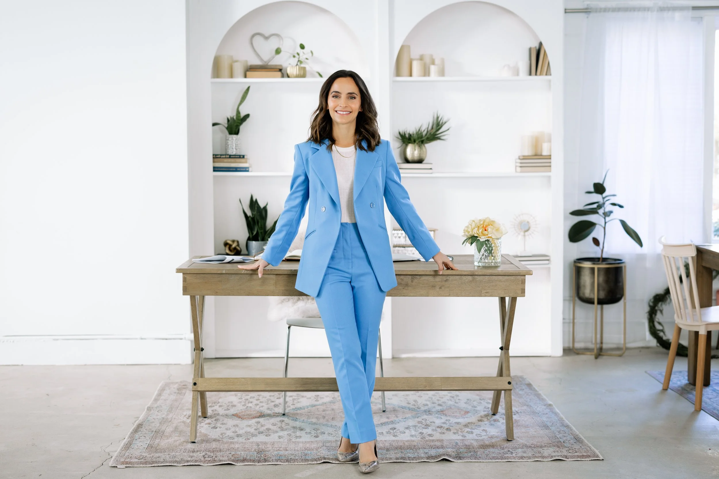 A brunette woman is leaning on a desk in a white office wearing a light blue suit