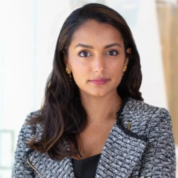 A woman with long dark hair, wearing a gray blazer with zip details, gold earrings, and makeup, looking directly at the camera.
