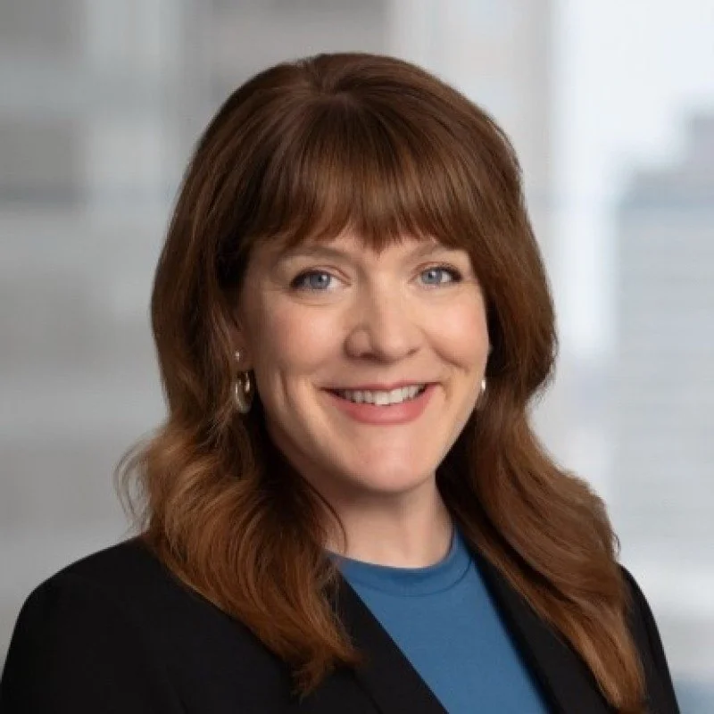A woman with shoulder-length red hair, wearing a black blazer and blue shirt, smiling in a professional environment.