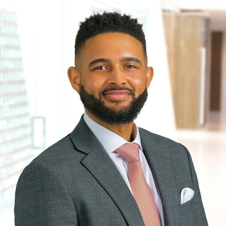 A man with a beard and short curly hair, dressed in a dark gray suit, light blue shirt, and pink tie, smiling in an office setting with blurred background.