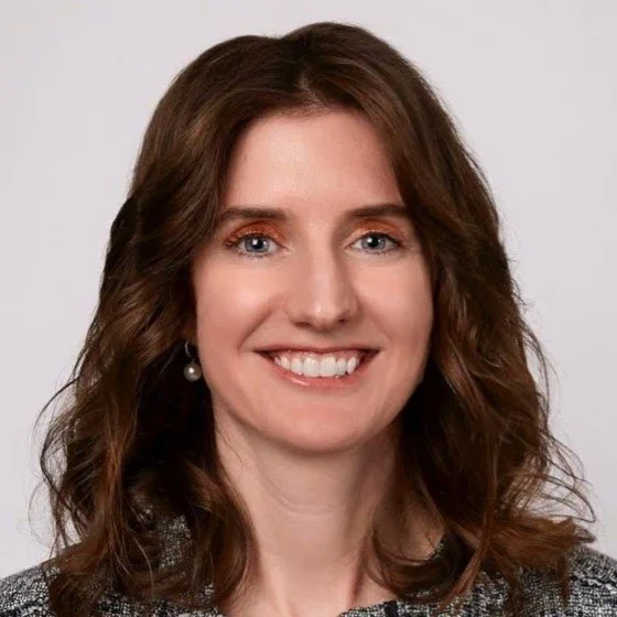 Close-up portrait of a woman with shoulder-length wavy brown hair, blue eyes, and fair skin, smiling and wearing a gray, textured top and pearl earrings against a plain light gray background.
