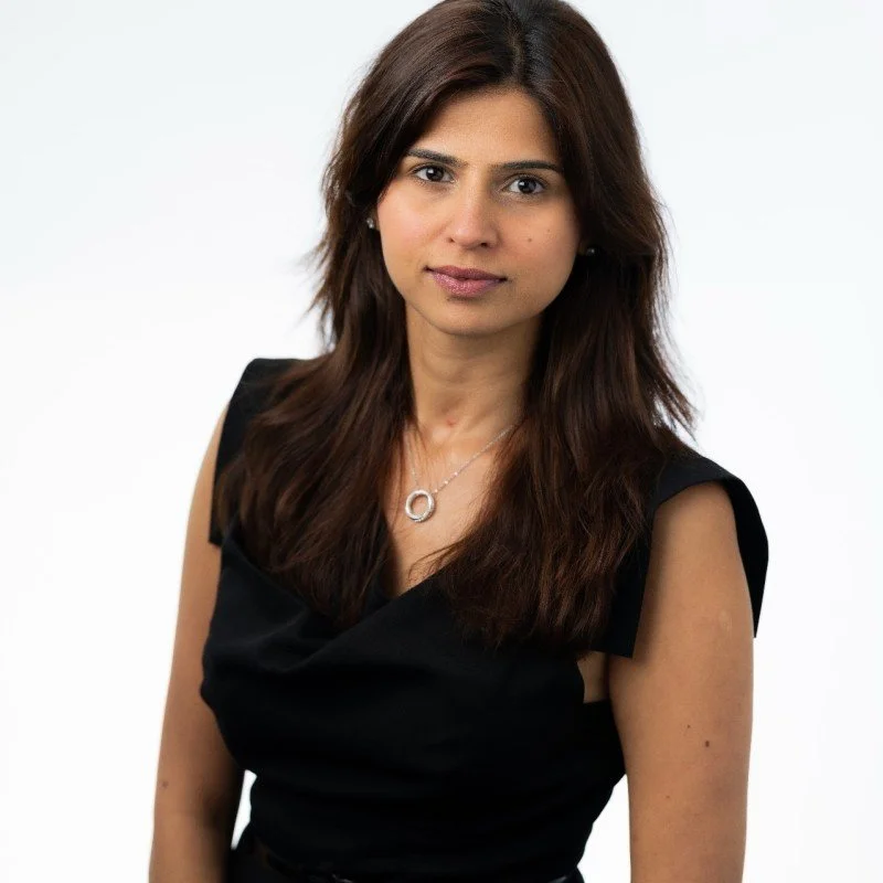 A woman with long brown hair wearing a black sleeveless top and a silver necklace, standing against a plain white background.