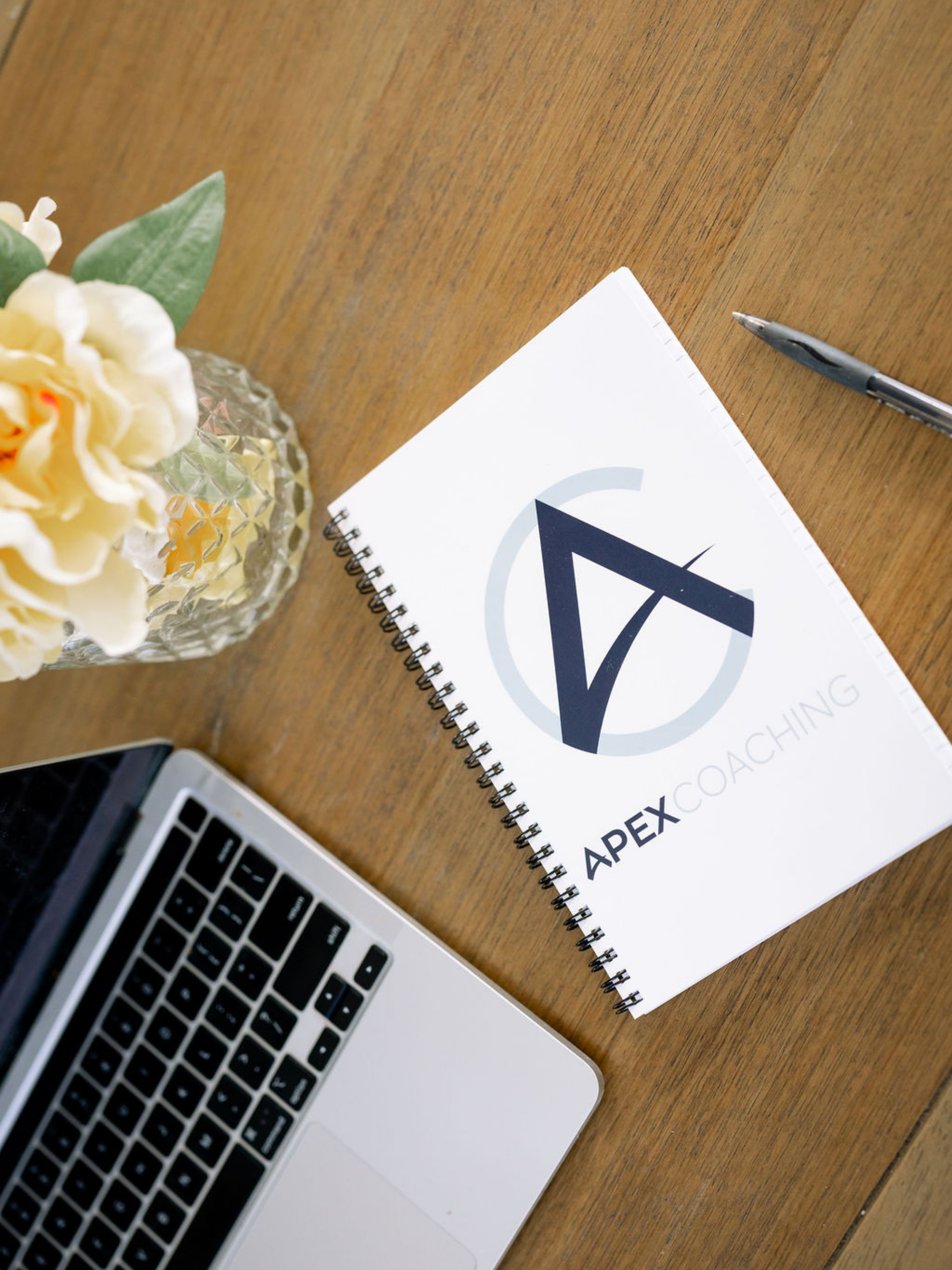 A laptop and notebook with Apex Coaching logo on a desk with flowers