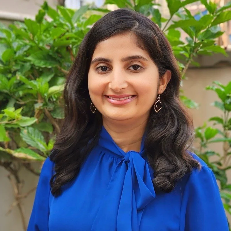 A woman with long dark hair smiling, wearing a blue blouse with a bow tie, standing outdoors with green leafy plants in the background.