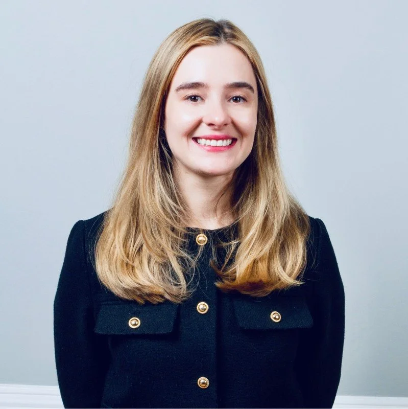 A young woman with long blonde hair, smiling, wearing a black jacket with gold buttons, standing against a light gray background.
