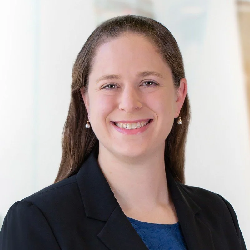 A smiling woman with light skin, long brown hair, wearing pearl earrings, a black blazer, and a blue top, standing indoors with a light background.