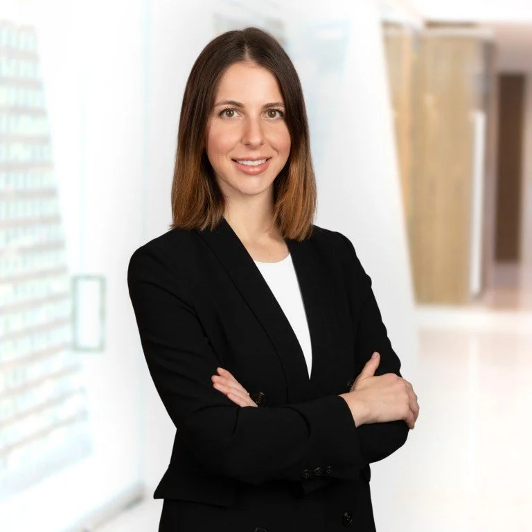 A woman with shoulder-length brown hair, wearing a black blazer over a white shirt, smiling with arms crossed in a bright office setting.