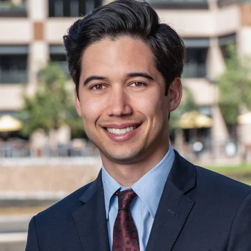 Young man in a suit smiling outdoors with buildings and trees in the background.