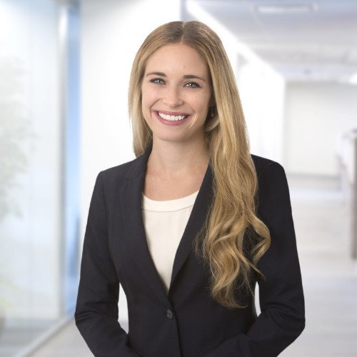 Professional woman with long blonde hair, wearing a black blazer and white top, standing in a modern office with smiling expression.