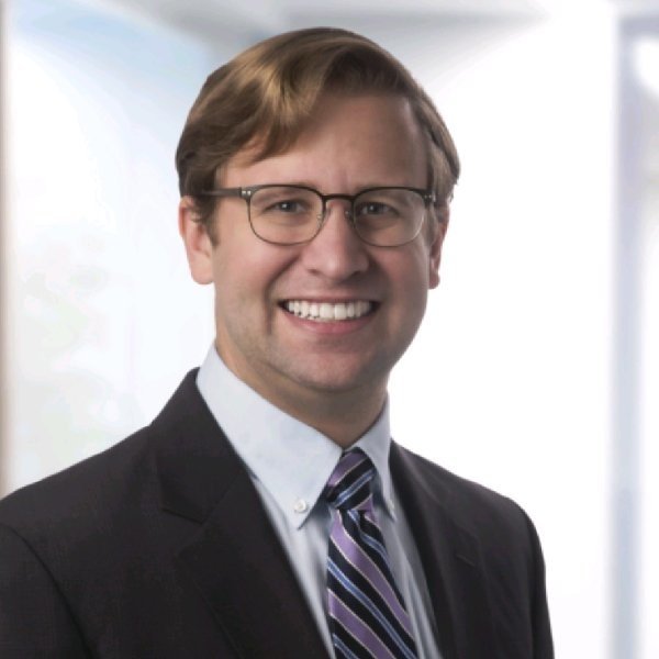 Portrait of a man wearing glasses, a suit, and a tie, smiling in a professional setting.