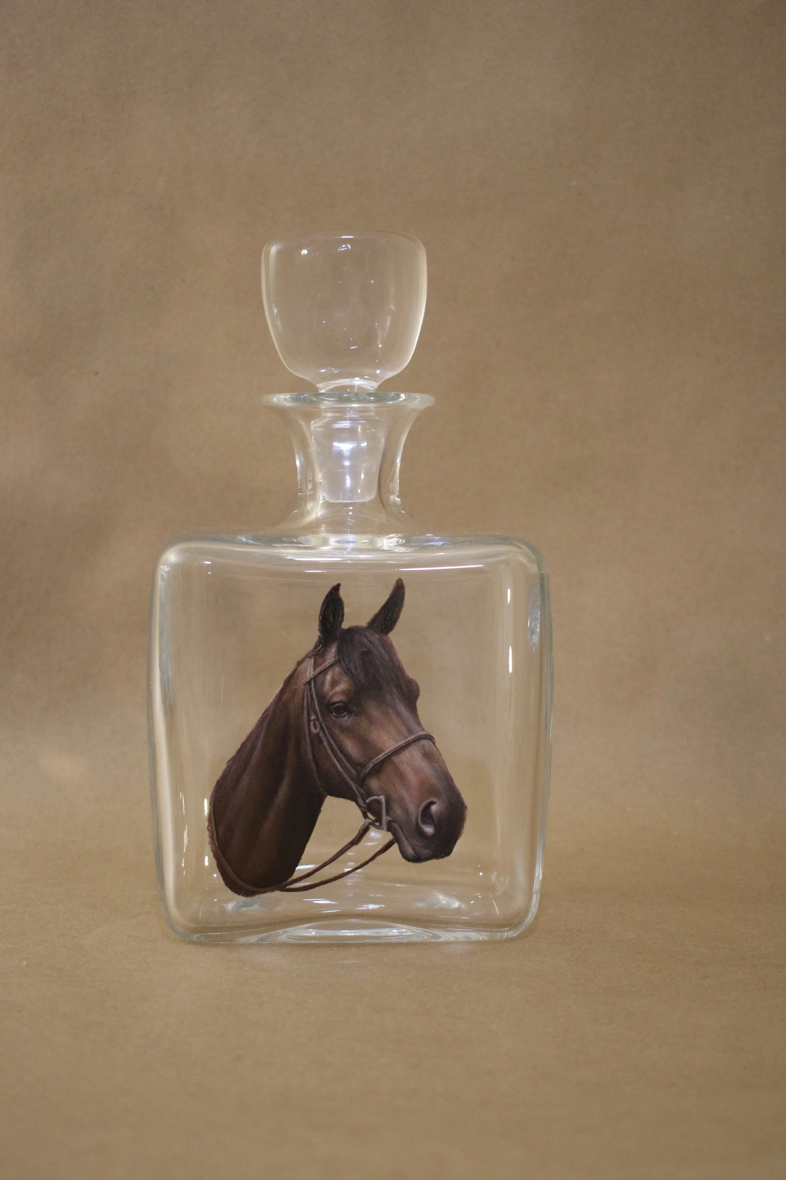 A glass decanter with a horse's head on the front and a glass stopper on top, set against a plain background.