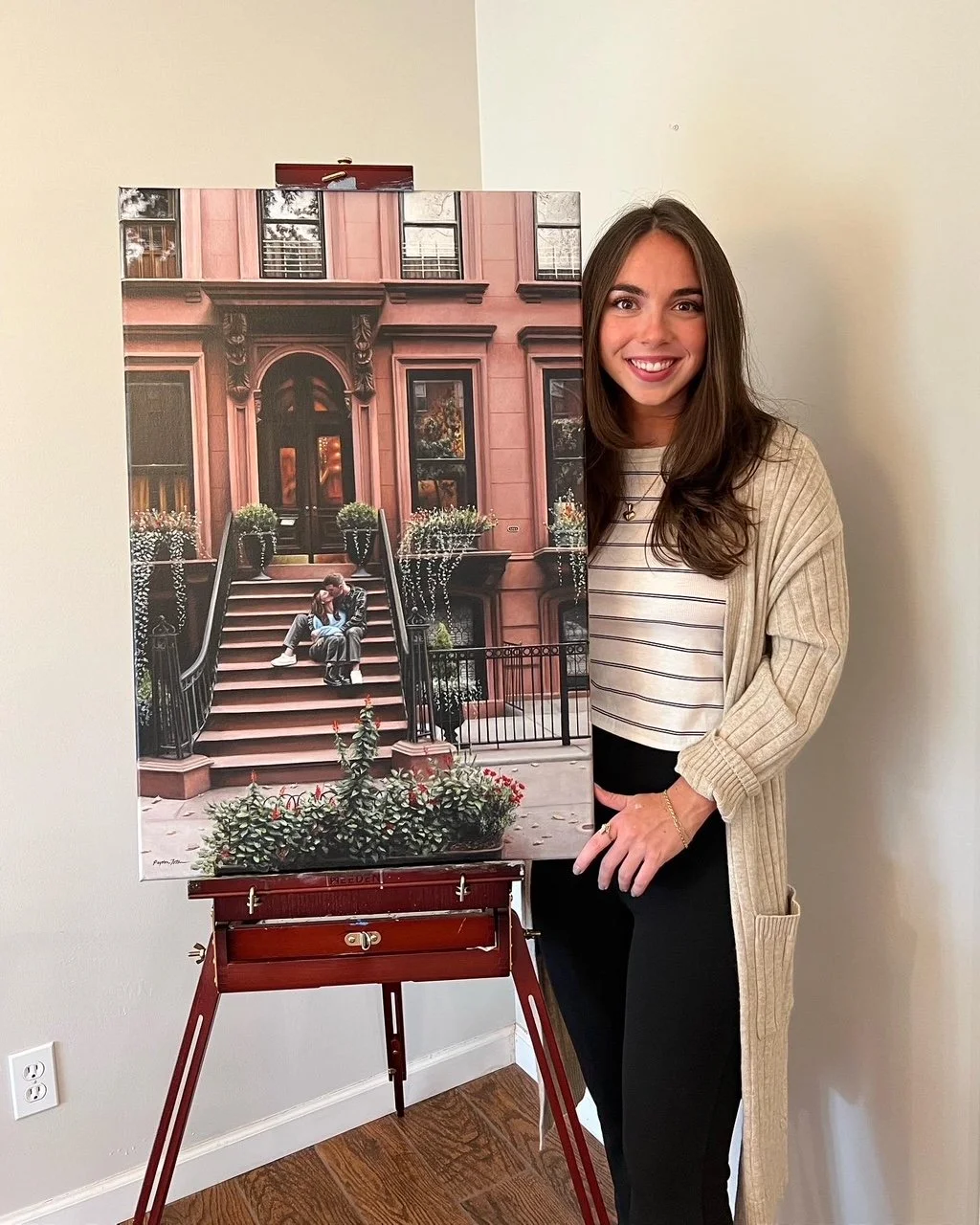 Woman in a beige cardigan standing next to a large painting of a brownstone building with people sitting on the steps.