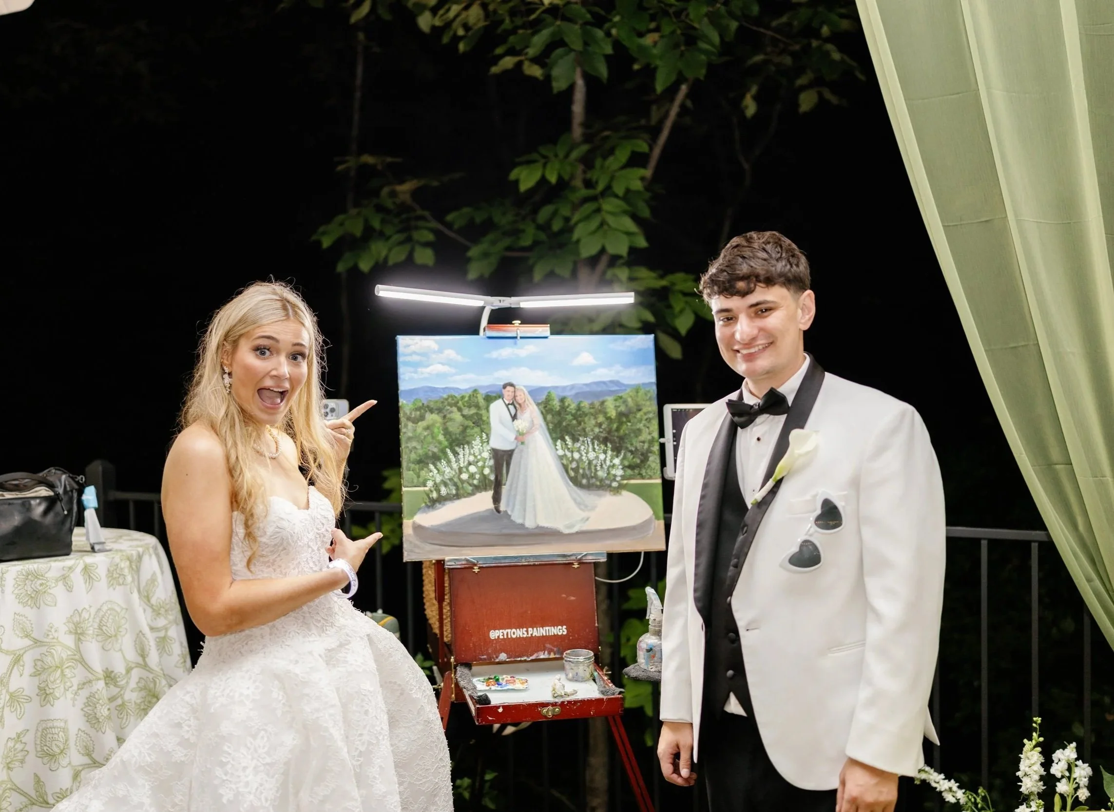 A woman in a wedding dress pointing excitedly at a live wedding painting of a bride and groom, with a man in a tuxedo smiling near the painting on a night outdoor setting.