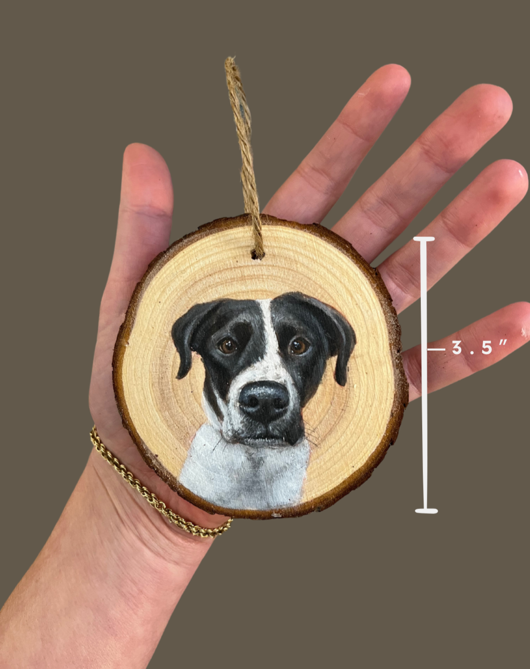 A hand holding a round wooden ornament with a painted portrait of a black and white dog. The ornament has a small hole at the top with a piece of string for hanging.
