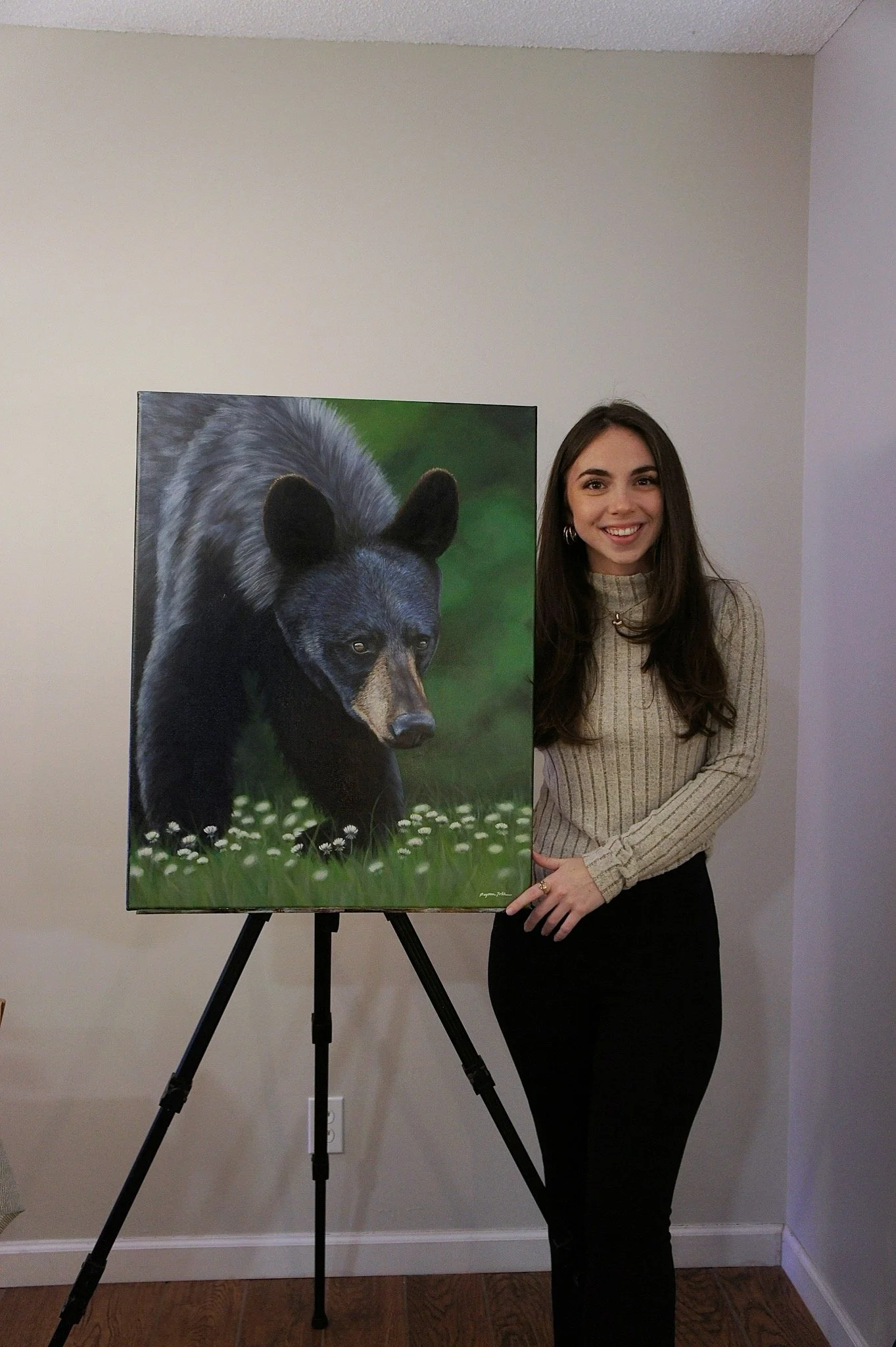 A woman standing next to a painting of a black bear walking on grass with small white flowers, indoors against a plain wall.