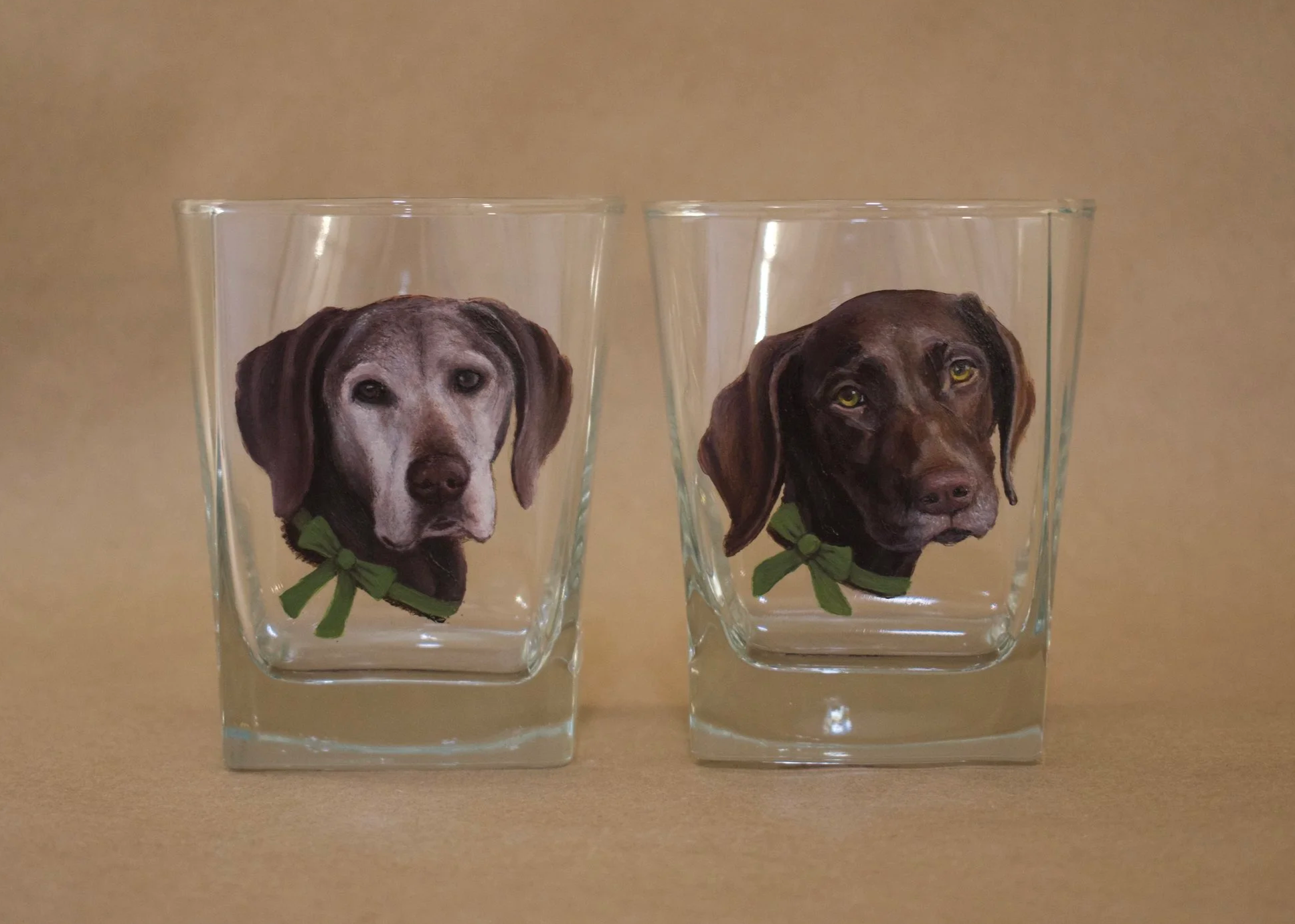 Two glass tumblers with illustrations of dogs wearing green collars, one with a gray design and the other with a brown design, placed on a tan background.