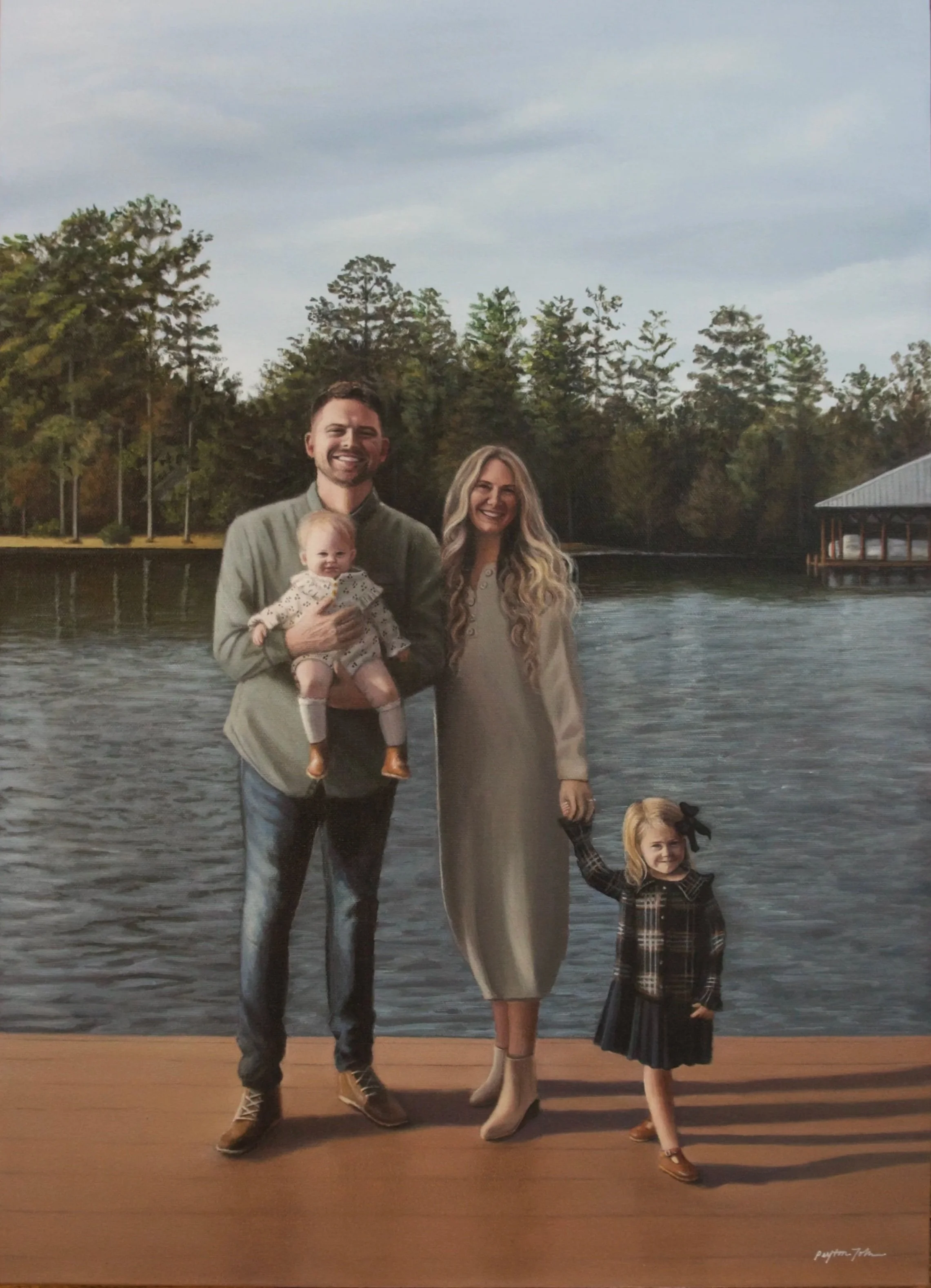 A family of four standing on a wooden dock by a lake with trees and a boathouse in the background, smiling at the camera.