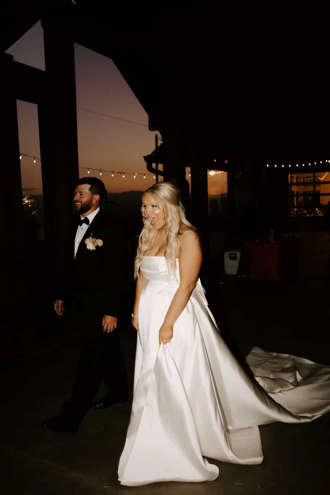 Bride in a white wedding gown and groom in a black tuxedo walking together during sunset at an outdoor wedding reception. Going to view their wedding painting