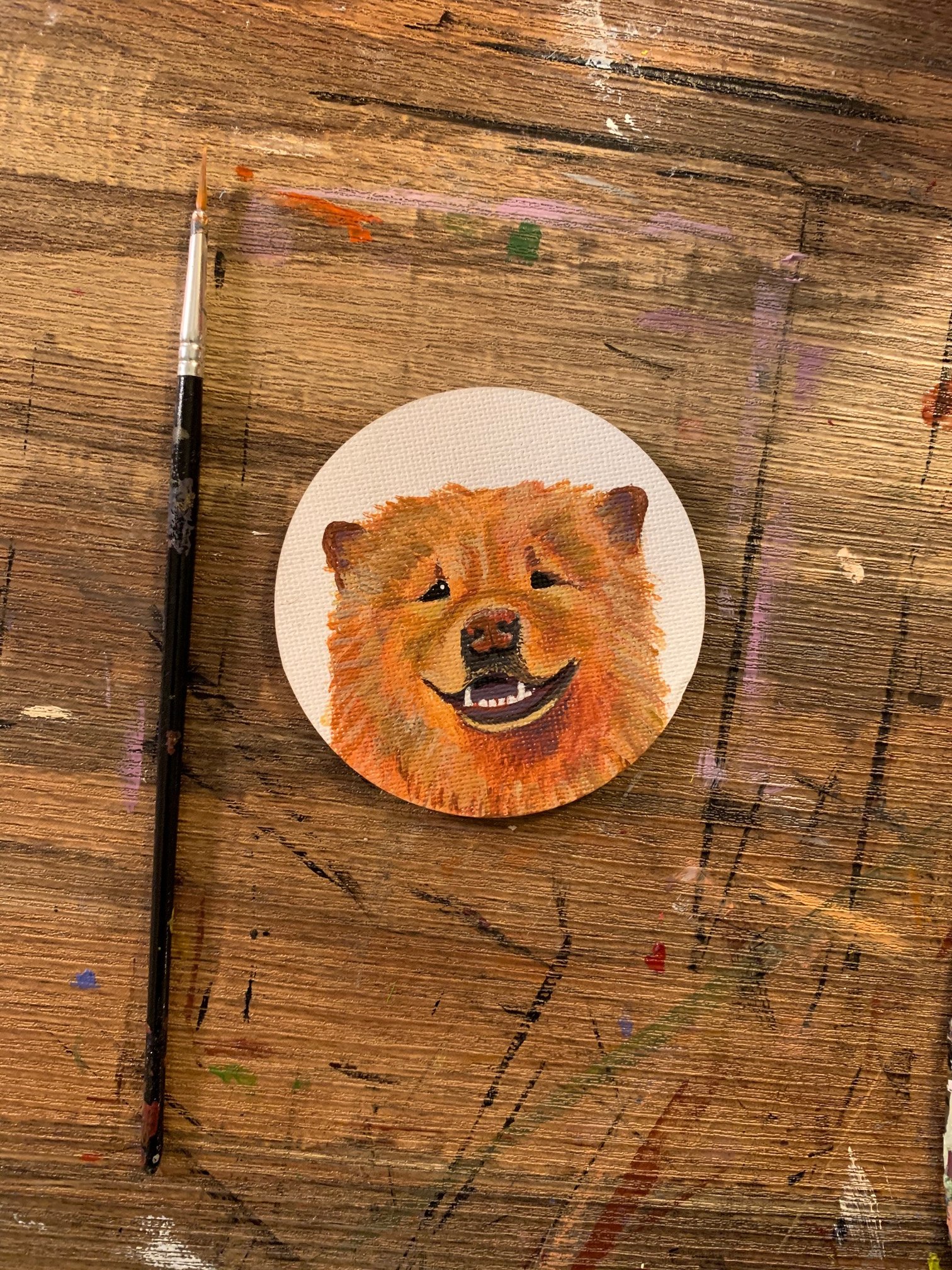 A small circular canvas with a watercolor painting of a happy, smiling dog with fluffy orange fur, on a wooden table with paint splatters and a paintbrush nearby.