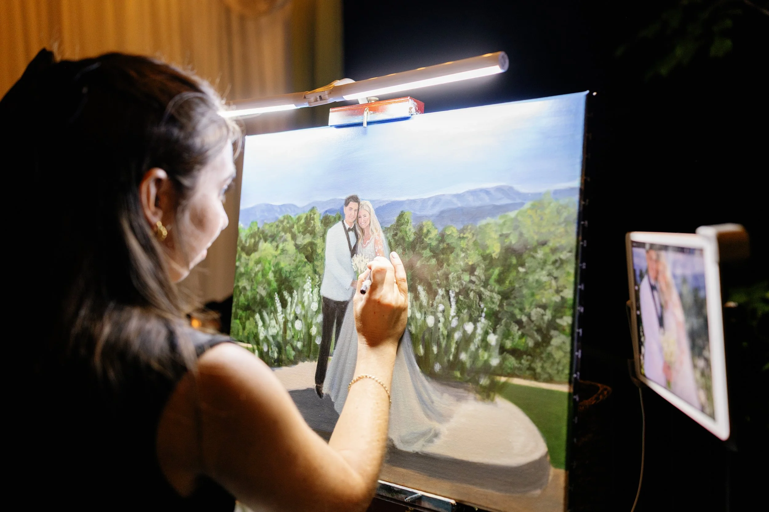 a live wedding painter artist painting a wedding portrait of a couple outdoors, with a landscape of trees and mountains in the background. The artist is a woman with dark hair, seen from the side, working at an easel with a light above.