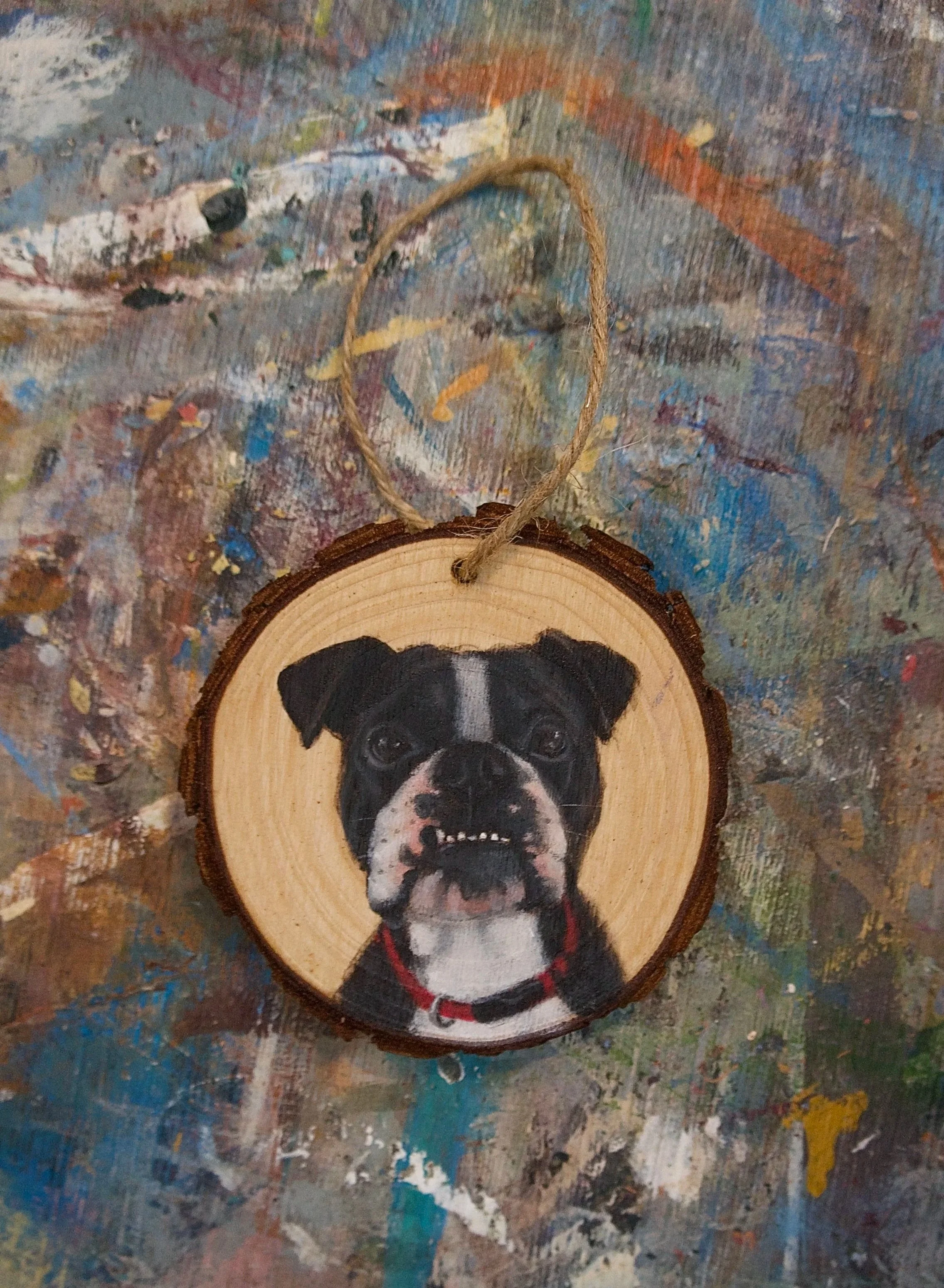 A circular wooden ornament featuring a painted portrait of a black and white Boston Terrier dog with a red collar, hanging by a thin string on a colorful, paint-splattered surface.