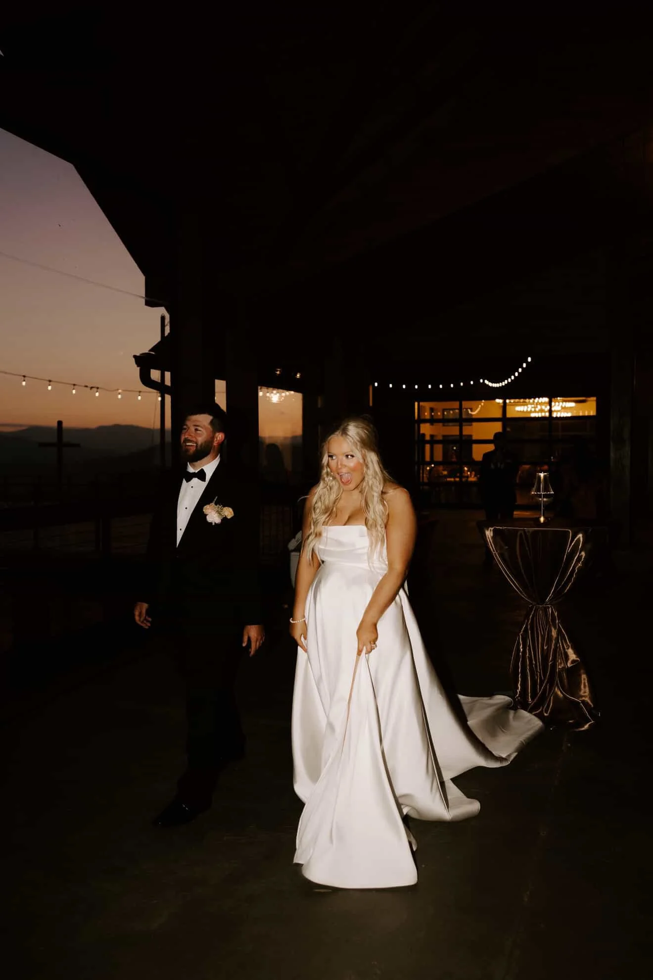 A bride in a white wedding dress and a groom in a black tuxedo with a bow tie walking together at sunset with outdoor string lights overhead.
