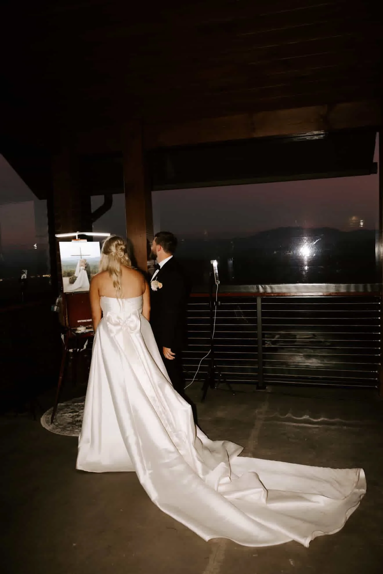 Bride and groom sharing a moment during their wedding reception at night, with mountains visible through a large window in the background. They are enjoying their new wedding painting created live at their wedding
