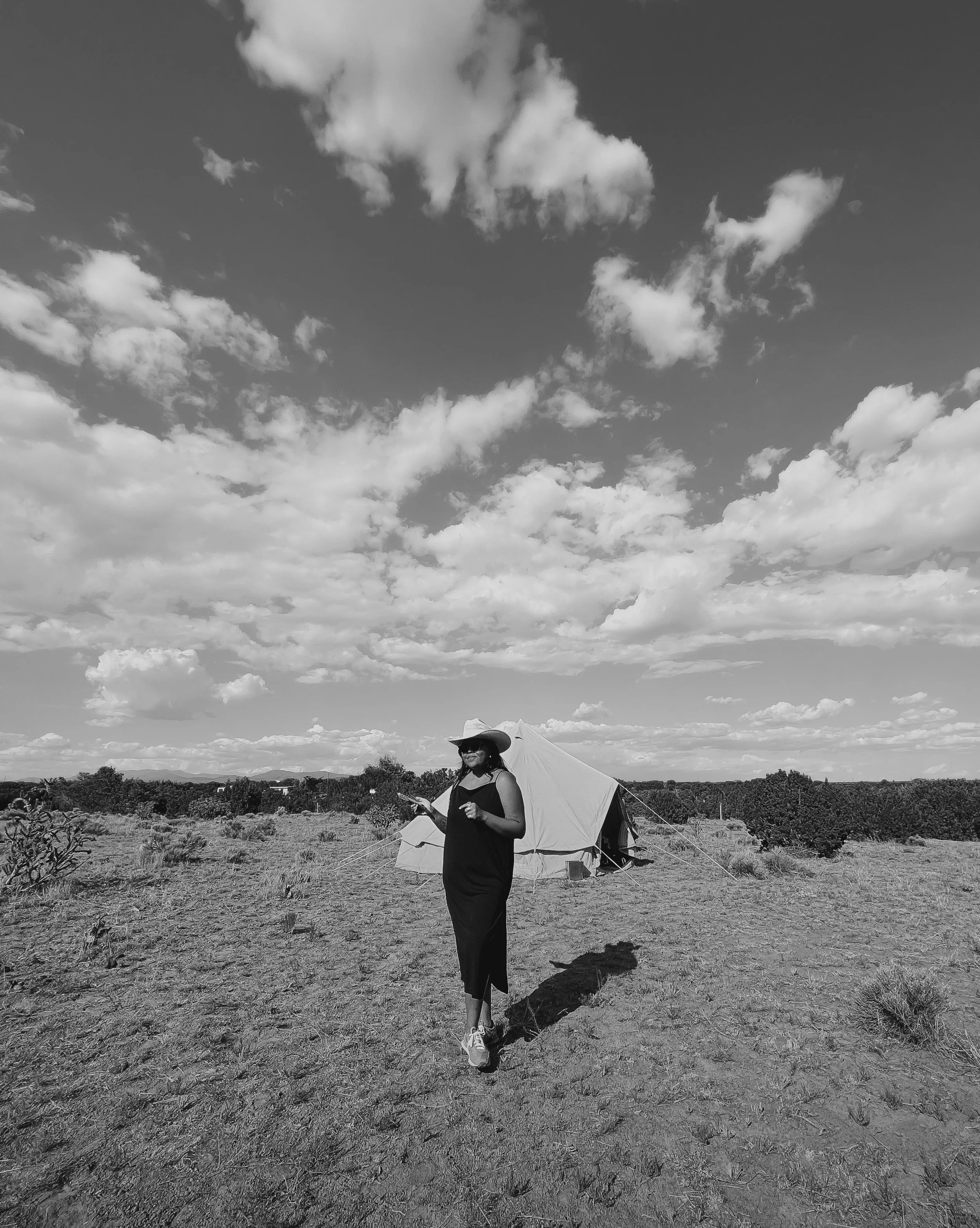 A woman in a black dress and hat standing outdoors in a field with a tent behind her, under a sky filled with clouds.