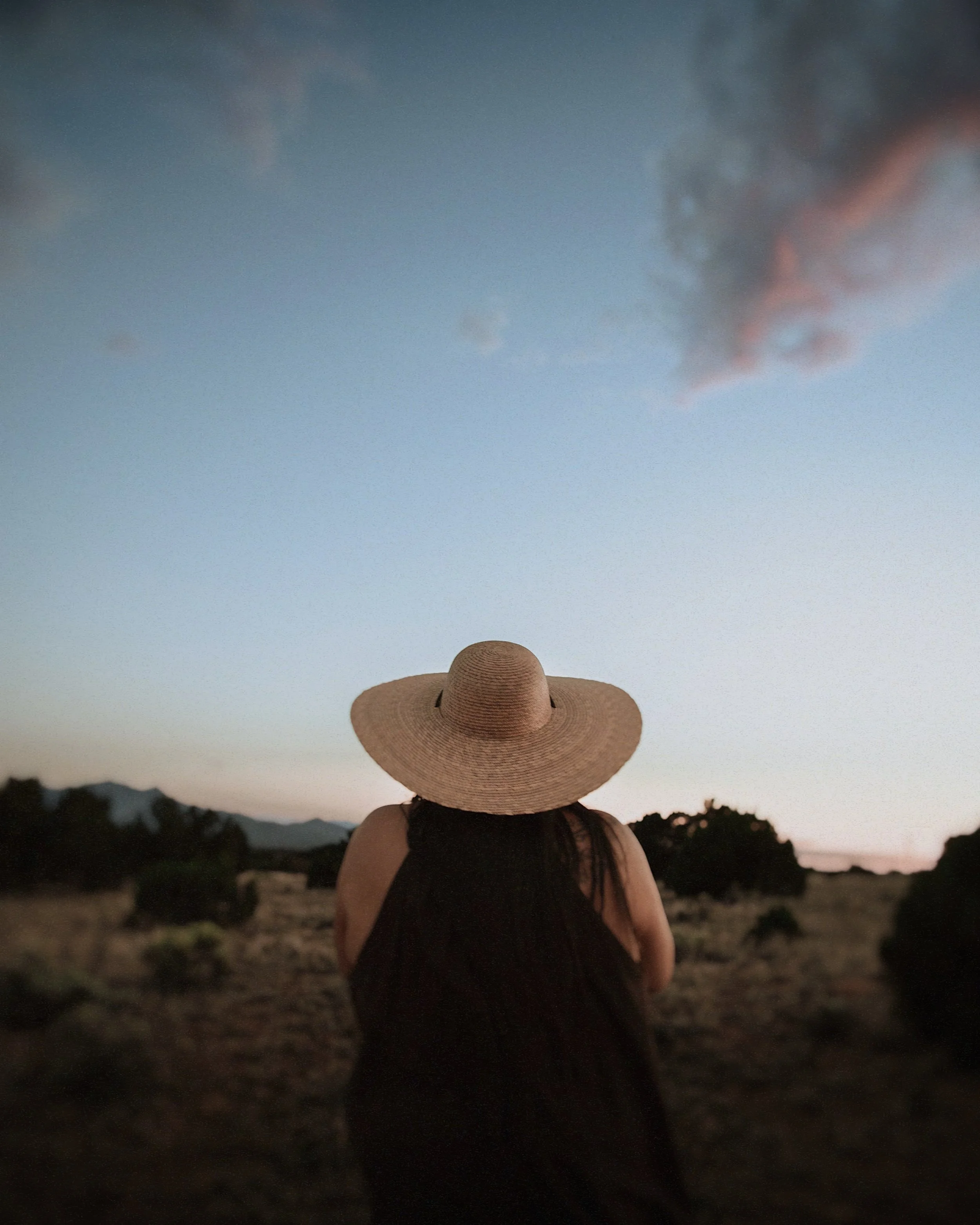 Woman with long dark hair wearing a wide-brimmed straw hat and black dress, standing in a field at dusk, looking at the sky with dark clouds and mountain silhouettes in the background.