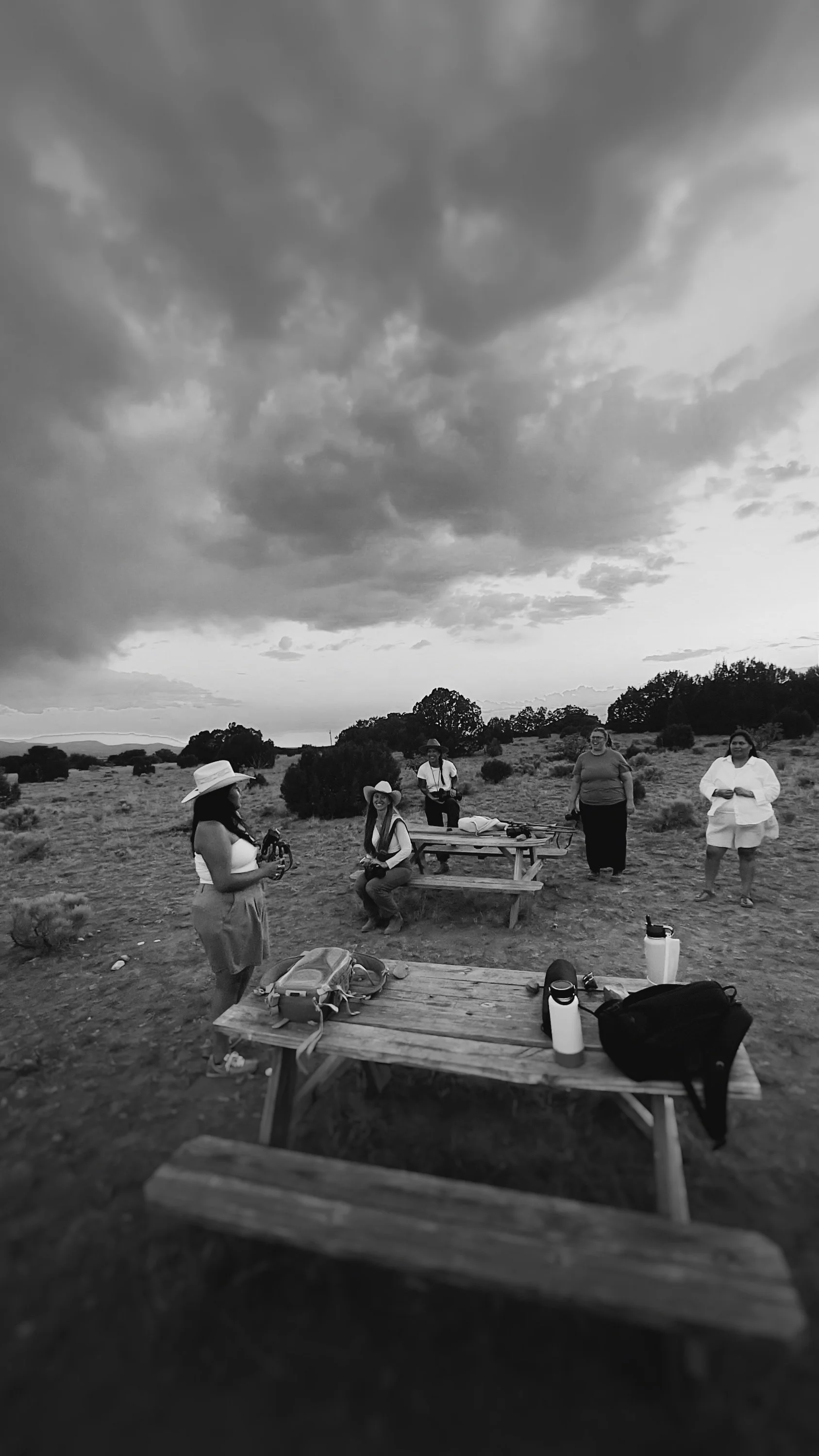 Group of people outdoors on a cloudy day with picnic tables and gear, wearing hats and casual clothing.