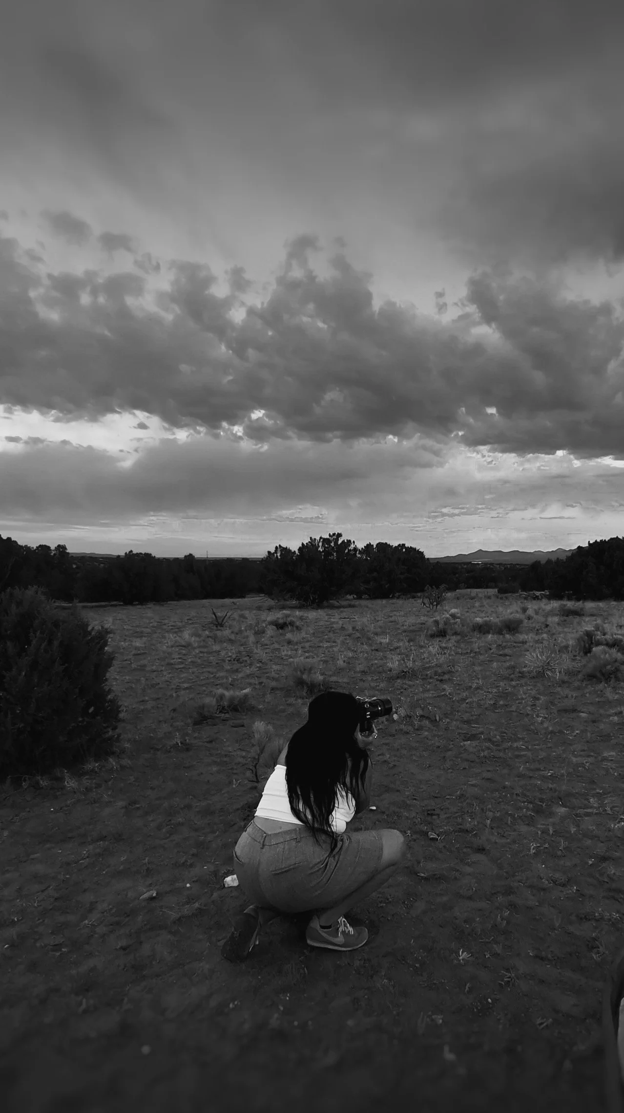 A woman crouching outdoors on dirt ground, holding a camera to her eye, aiming at the sky and landscape. She has long dark hair, wears a white top, shorts, and sneakers. The background features an open field with sparse bushes, trees, distant mountai