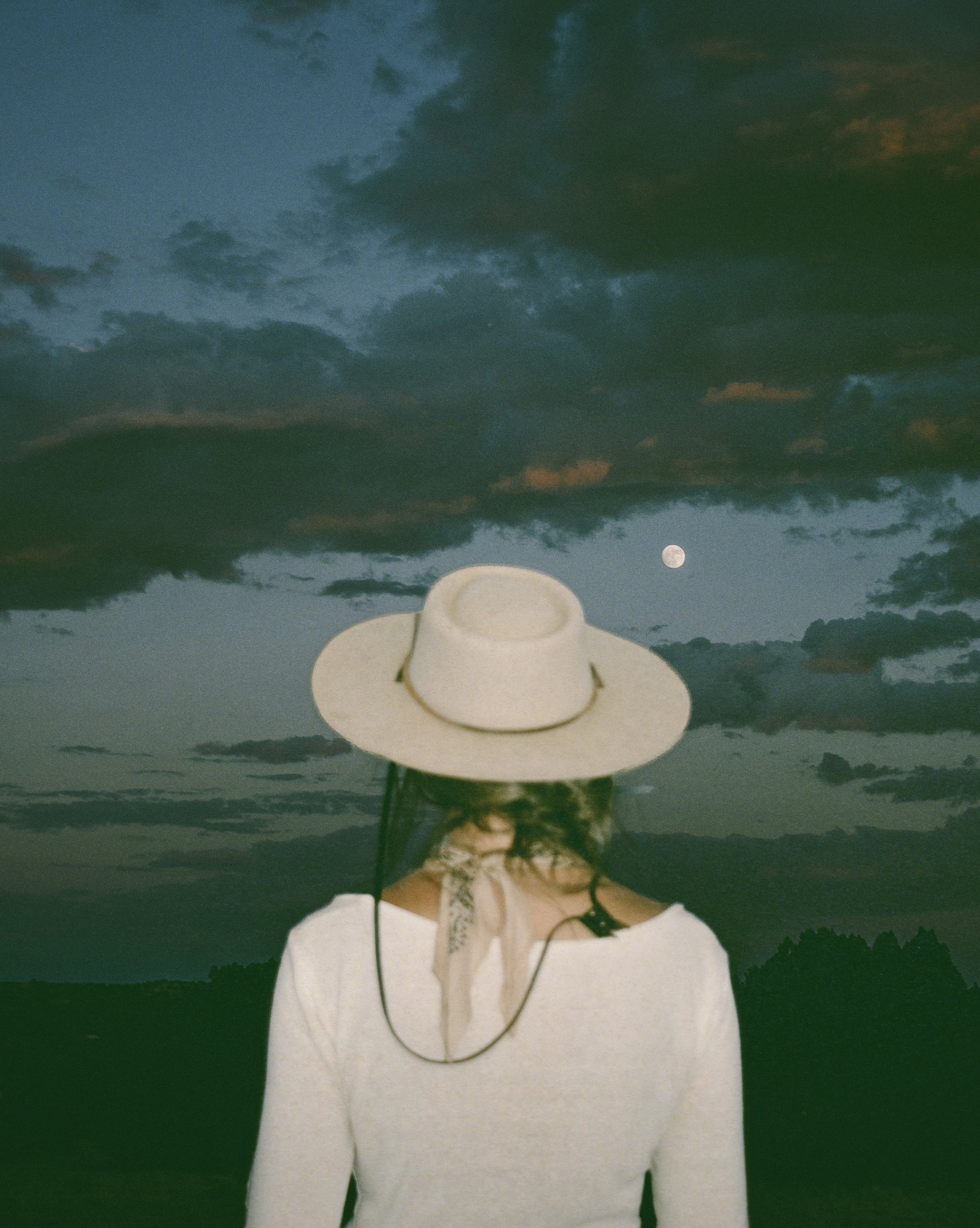 A person wearing a wide-brimmed hat and a light-colored top faces away, with a night sky, clouds, and the moon in the background.