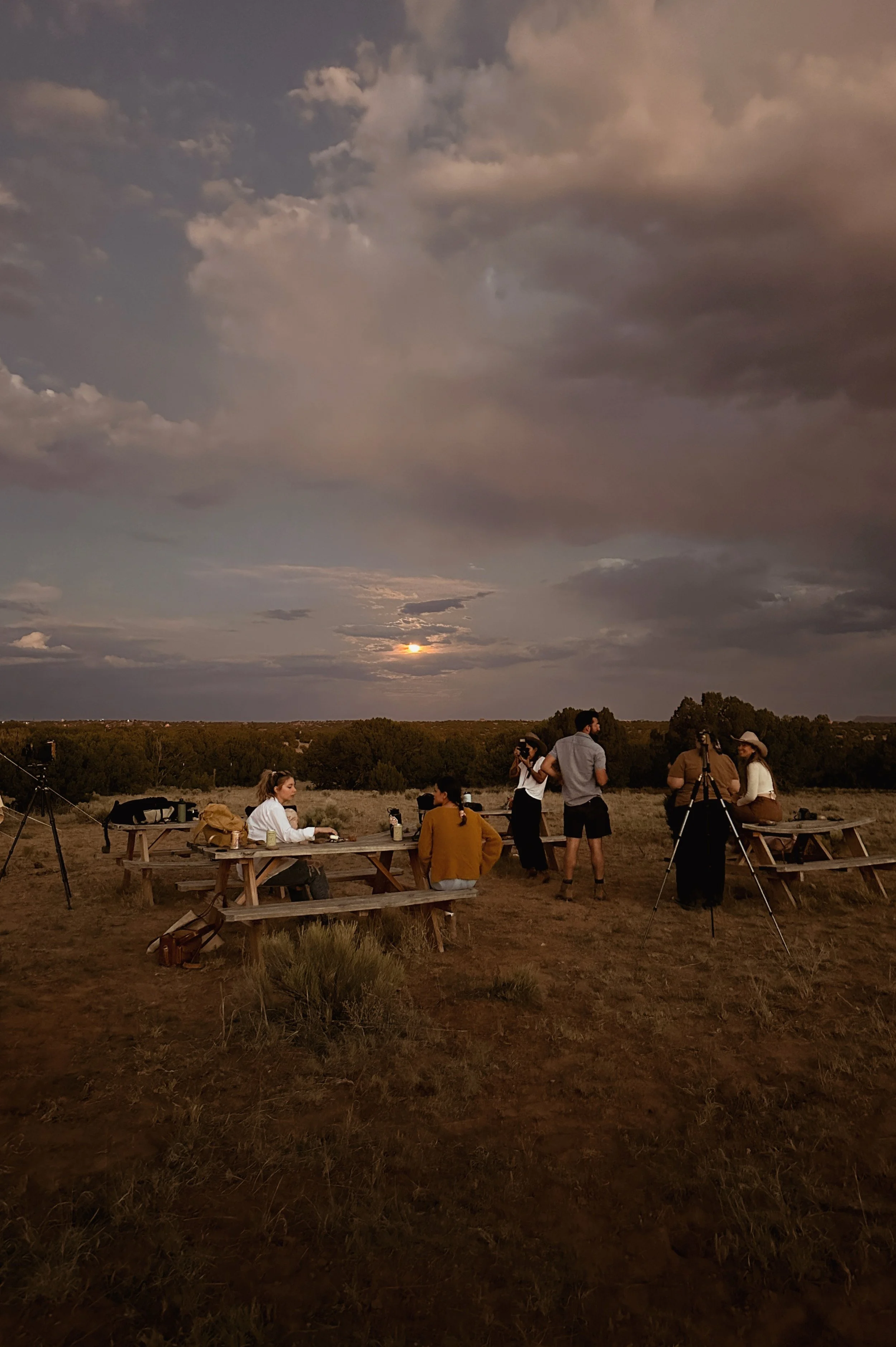 A group of people gathered outdoors during sunset, with some sitting at picnic tables and others standing or setting up cameras for star or moon photography in a rural field with trees in the background.