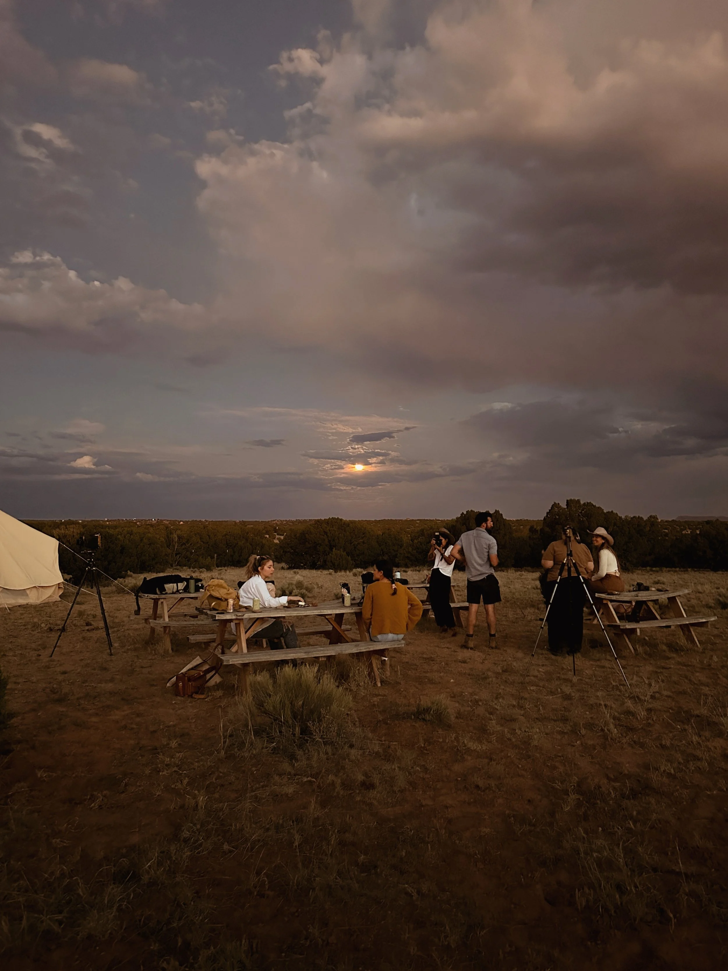 Group of people gathered outdoors at sunset, some sitting at picnic tables and others standing around with cameras and tripods, in a natural landscape with a cloudy sky and a setting sun.
