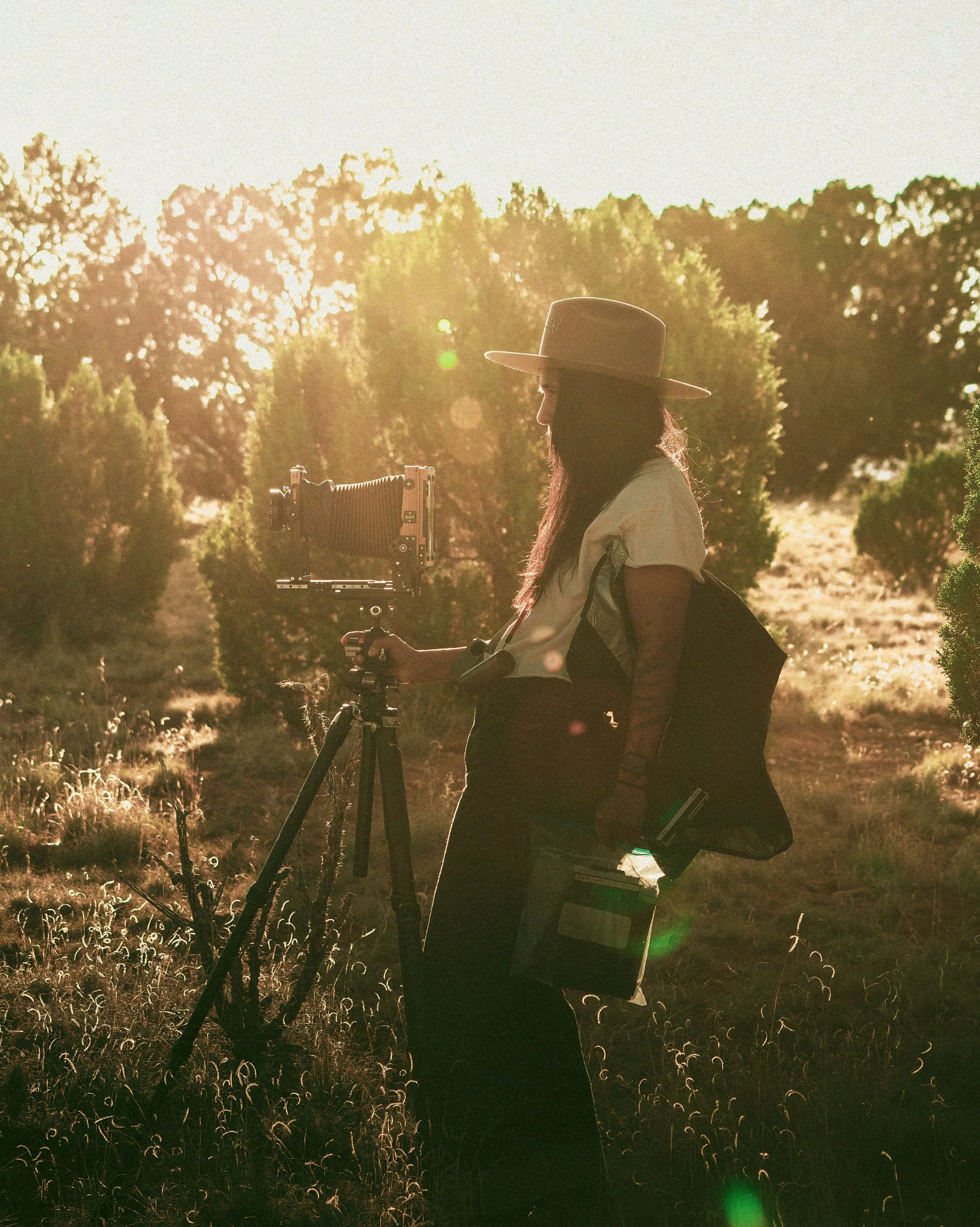 A woman in a wide-brimmed hat standing outdoors in a field with trees, holding a vintage camera on a tripod, backlit by the setting sun.