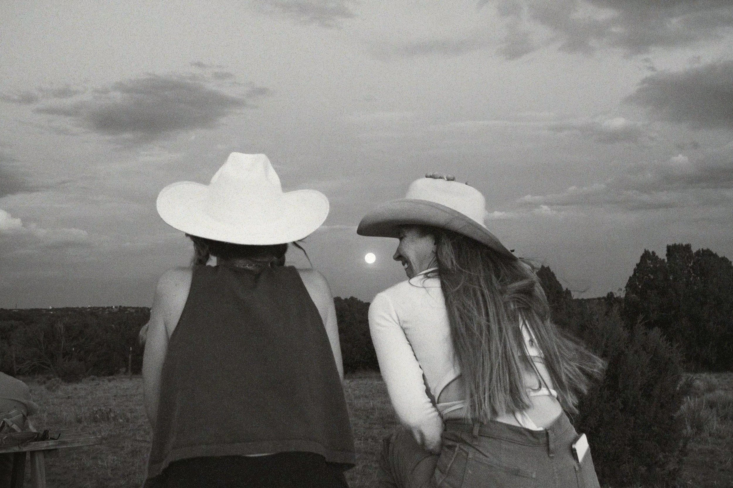 Two women wearing cowboy hats sitting outdoors at dusk, smiling and looking at each other, with the moon rising behind them and cloudy sky overhead.