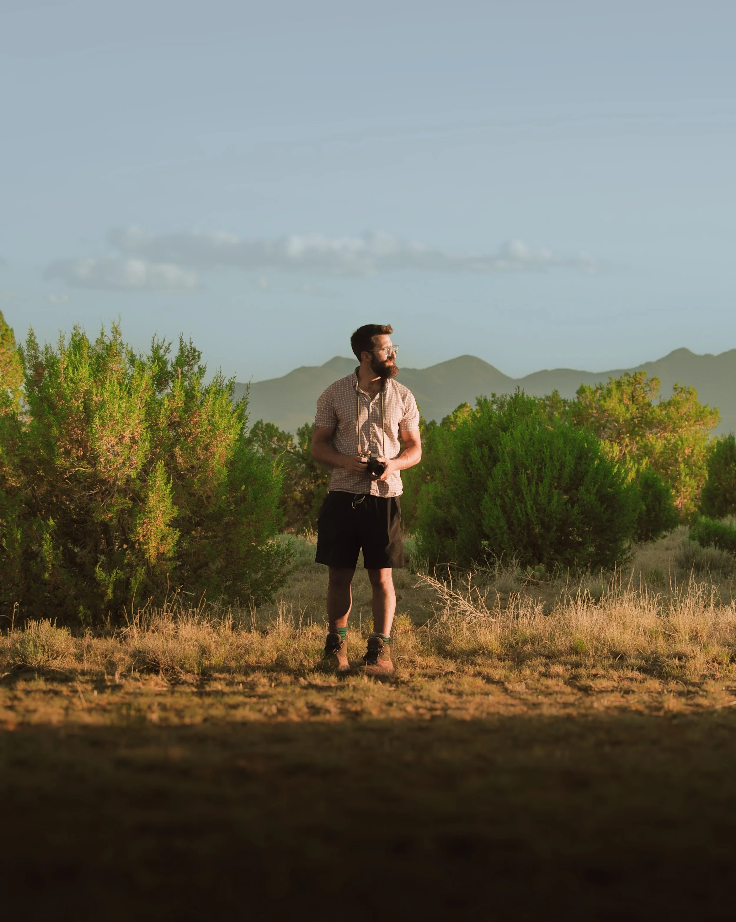 A man standing outdoors in a field holding a camera, with trees and mountains in the background, during sunset.