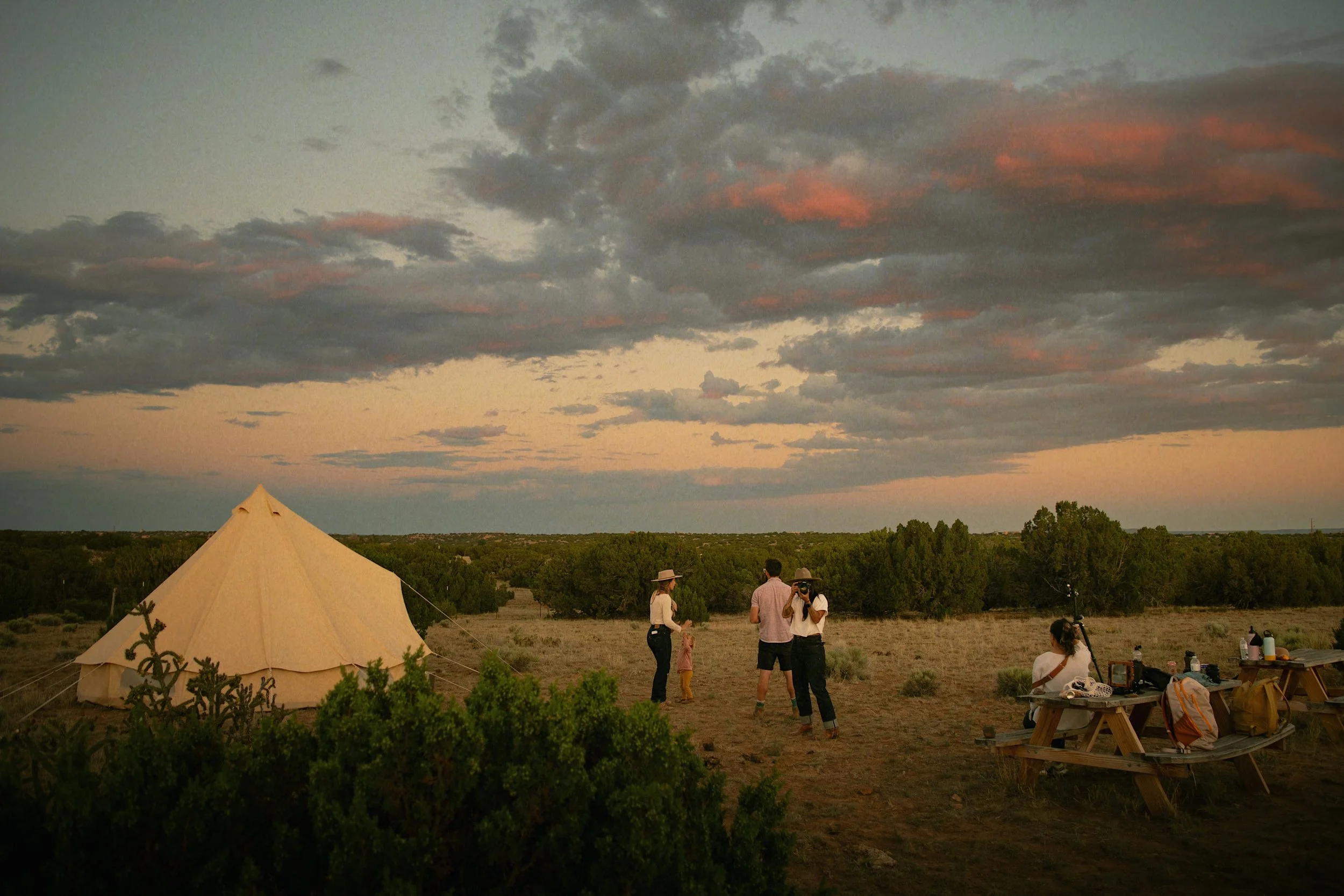 A group of people camping outdoors during sunset with a large beige teepee tent, trees in the background, and some picnic tables with belongings.